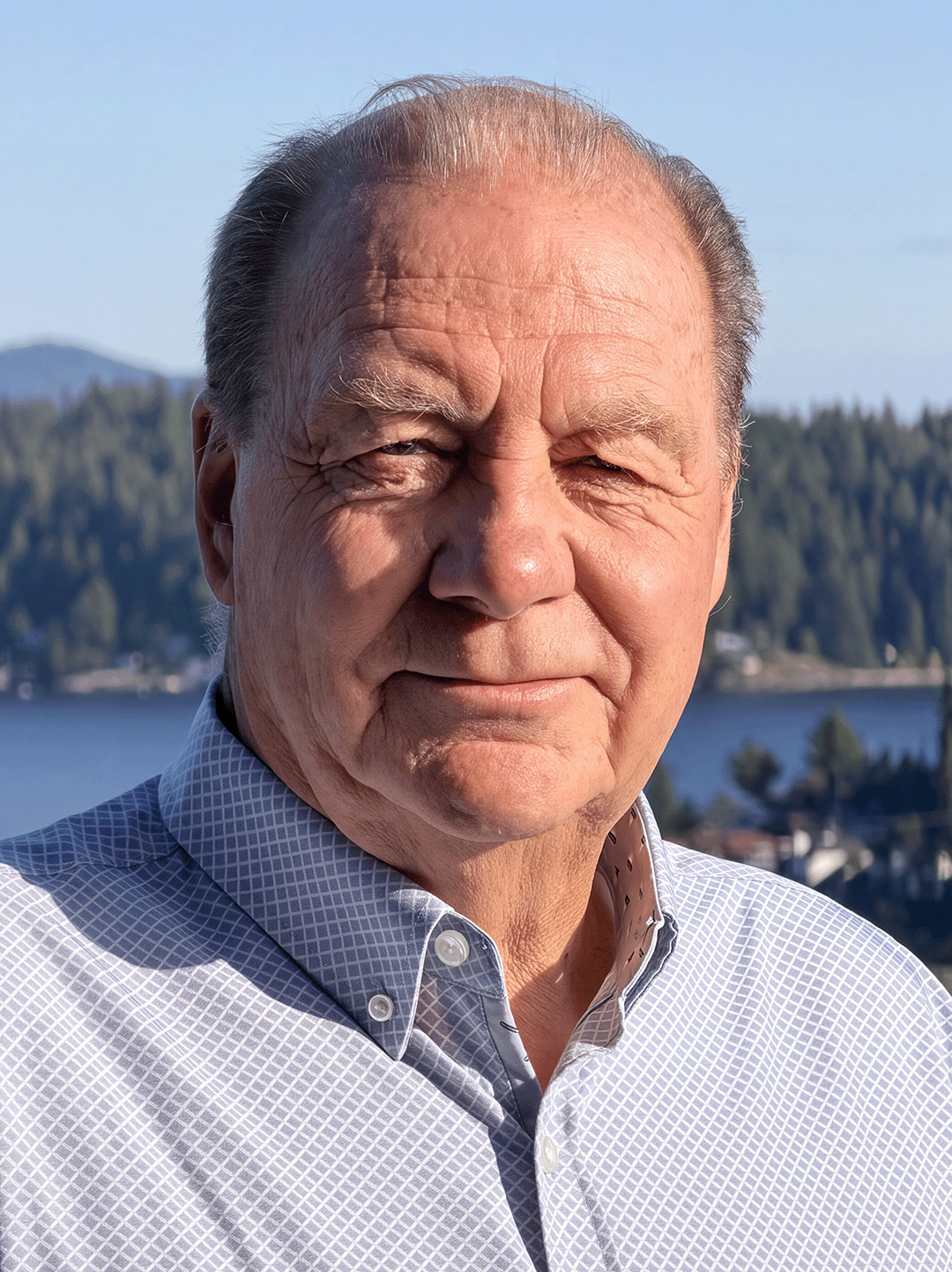Des Delaney, smiling outdoors with the Gibsons harbour and forest in the background
